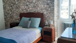 Une chambre avec un mur en pierre, un lit doté d’une tête de lit en bois, une table de chevet et une fenêtre donnant sur des immeubles.