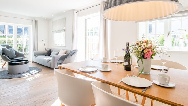 Una sala de estar moderna con una mesa de comedor de madera preparada para dos personas, un sillón gris, un sofá gris y un amplio ventanal que proporciona luz natural.