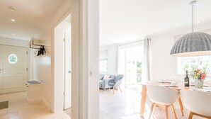 Un interior luminoso y moderno con suelo de madera, una zona de comedor con sillas blancas y una mesa, y un gran ventanal que deja entrar luz natural.