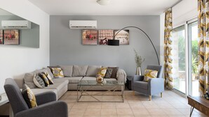 Una sala de estar moderna con un sofá modular, una mesa de centro de vidrio y un sillón gris. Se observan tres aires acondicionados instalados en la pared y un ventanal con cortinas estampadas.
