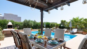 Comedor al aire libre con vista a la piscina, sillas de madera y una parrilla.