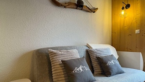 A cozy living room with a grey sofa, two grey pillows with mountain designs, a small white side table, and a wooden wall with a hanging light fixture. - 2-room cabin apartment for 4, south-facing terrace (Valloire)