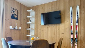 A wooden-paneled room with a round dining table, chairs, a flat-screen TV, and skis leaning against the wall. - 2-room cabin apartment for 4, south-facing terrace (Valloire)