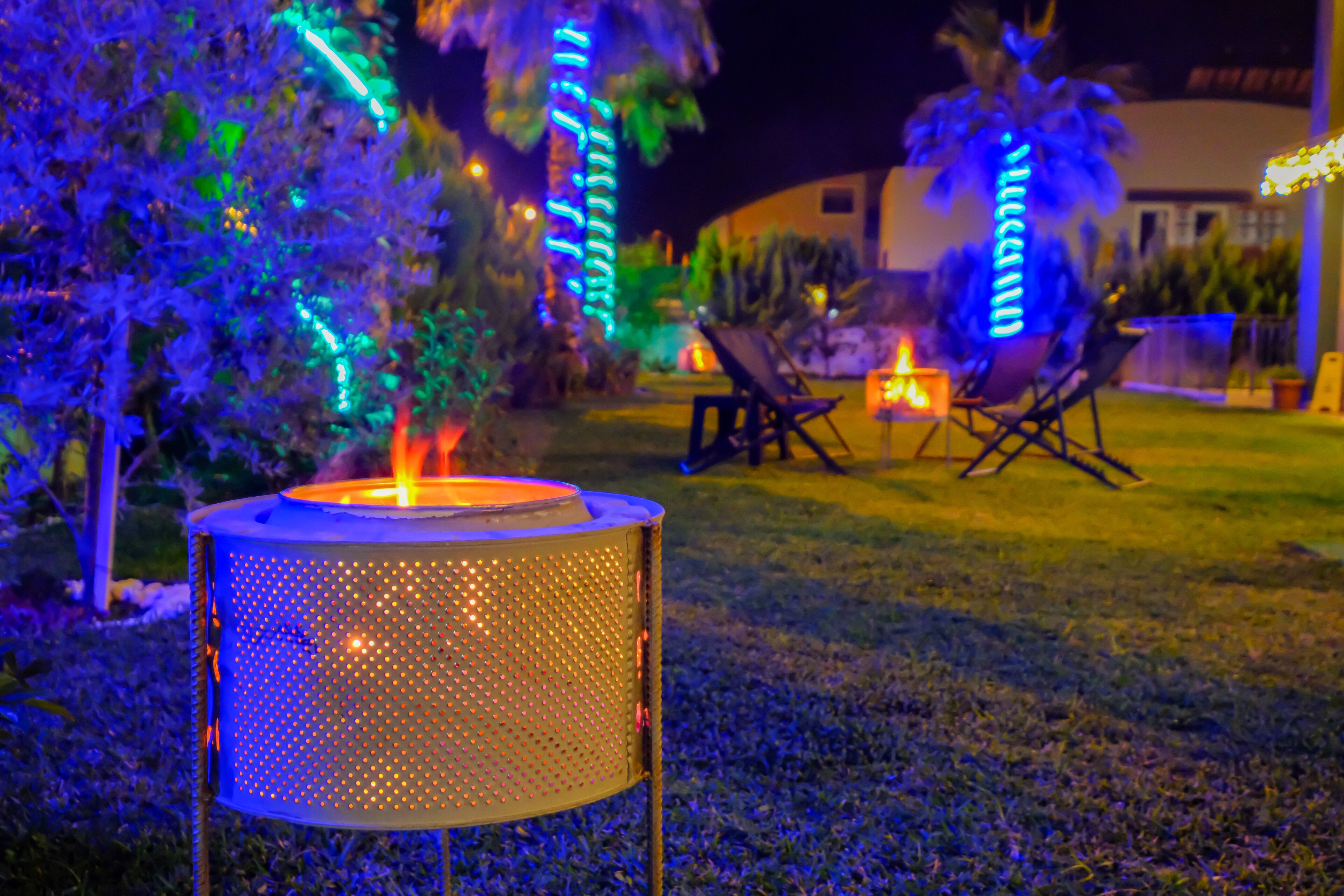 A metal grill with a lit burner in a garden at night, surrounded by trees and chairs.