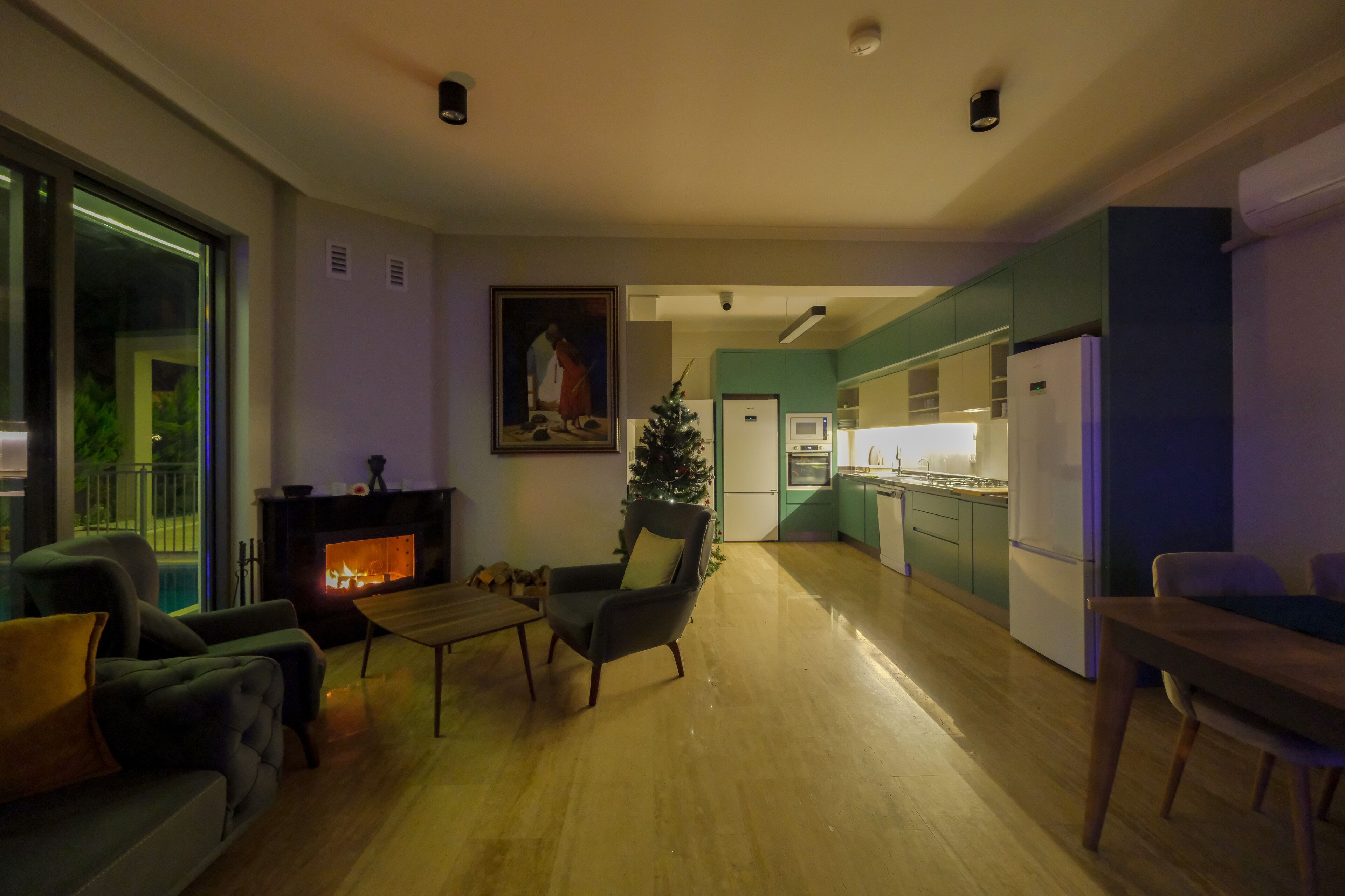 A modern living room with a fireplace, a dining area, and a kitchen with teal cabinets.