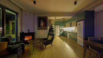 A modern living room with a fireplace, a dining area, and a kitchen with teal cabinets.