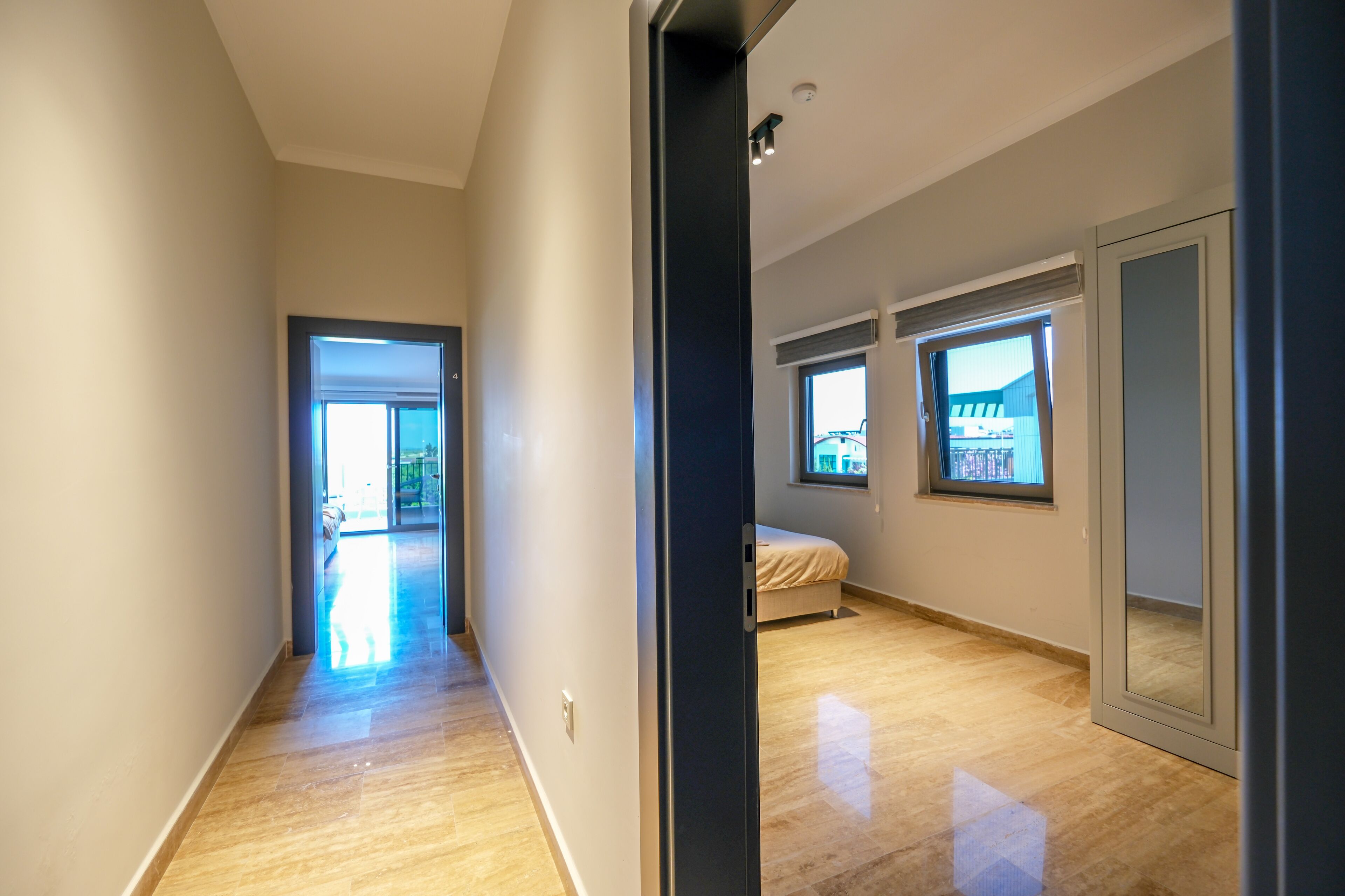 A modern, well-lit hallway leading to a bedroom with a large window, a door, and a visible balcony.