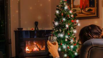 A person sitting by a fireplace with a decorated Christmas tree and a painting on the wall.