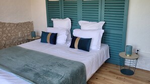 Una cama con ropa blanca y un cabecero verde azulado, una manta verde y una mesita redonda con un florero azul.