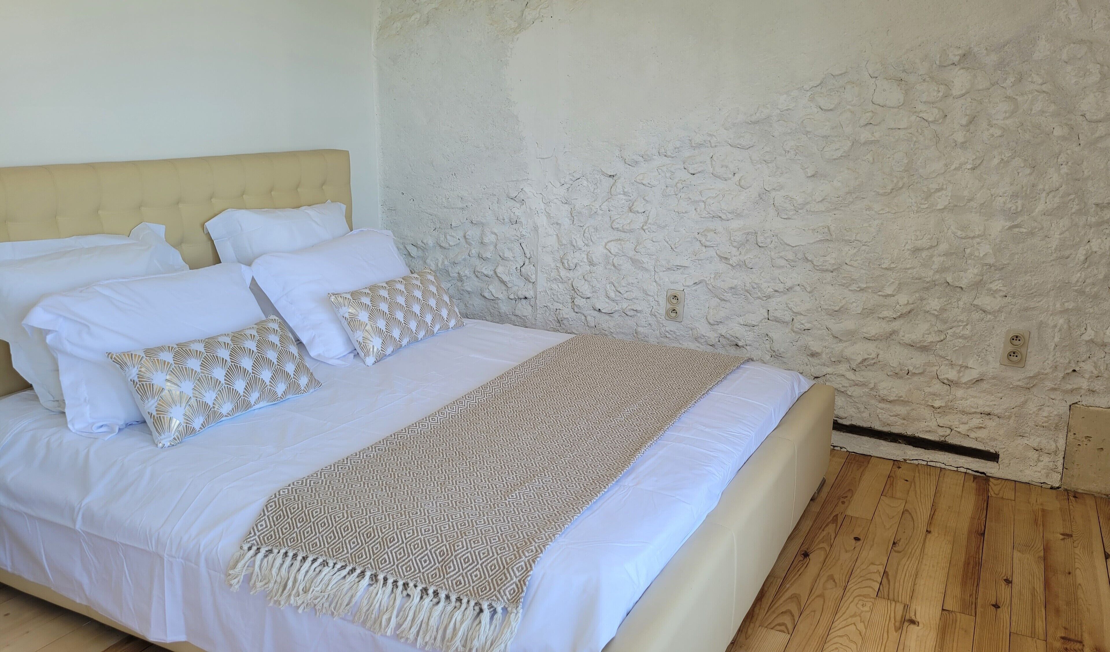 Un lit avec une literie blanche et des oreillers à motifs, un plaid beige et un mur texturé en arrière-plan.