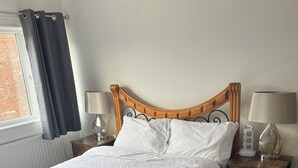 A neatly made bed with white linens, a wooden headboard, two bedside tables with lamps, a black rug, and a window with curtains. - Fellowes Garden (Peterborough)