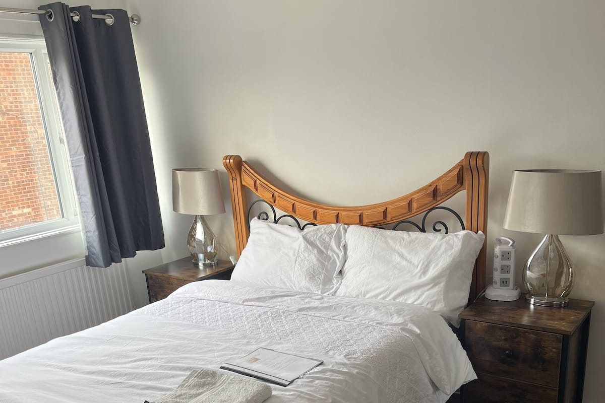 A neatly made bed with white linens, a wooden headboard, two bedside tables with lamps, a black rug, and a window with curtains.
