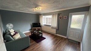 A living room with a grey sofa, a wooden coffee table, a TV on a stand, and a window with curtains. - Fellowes Garden (Peterborough)