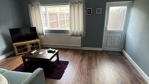 A living room with a flat-screen TV, wooden flooring, a sofa, a coffee table, and a window with curtains. - Fellowes Garden (Peterborough)
