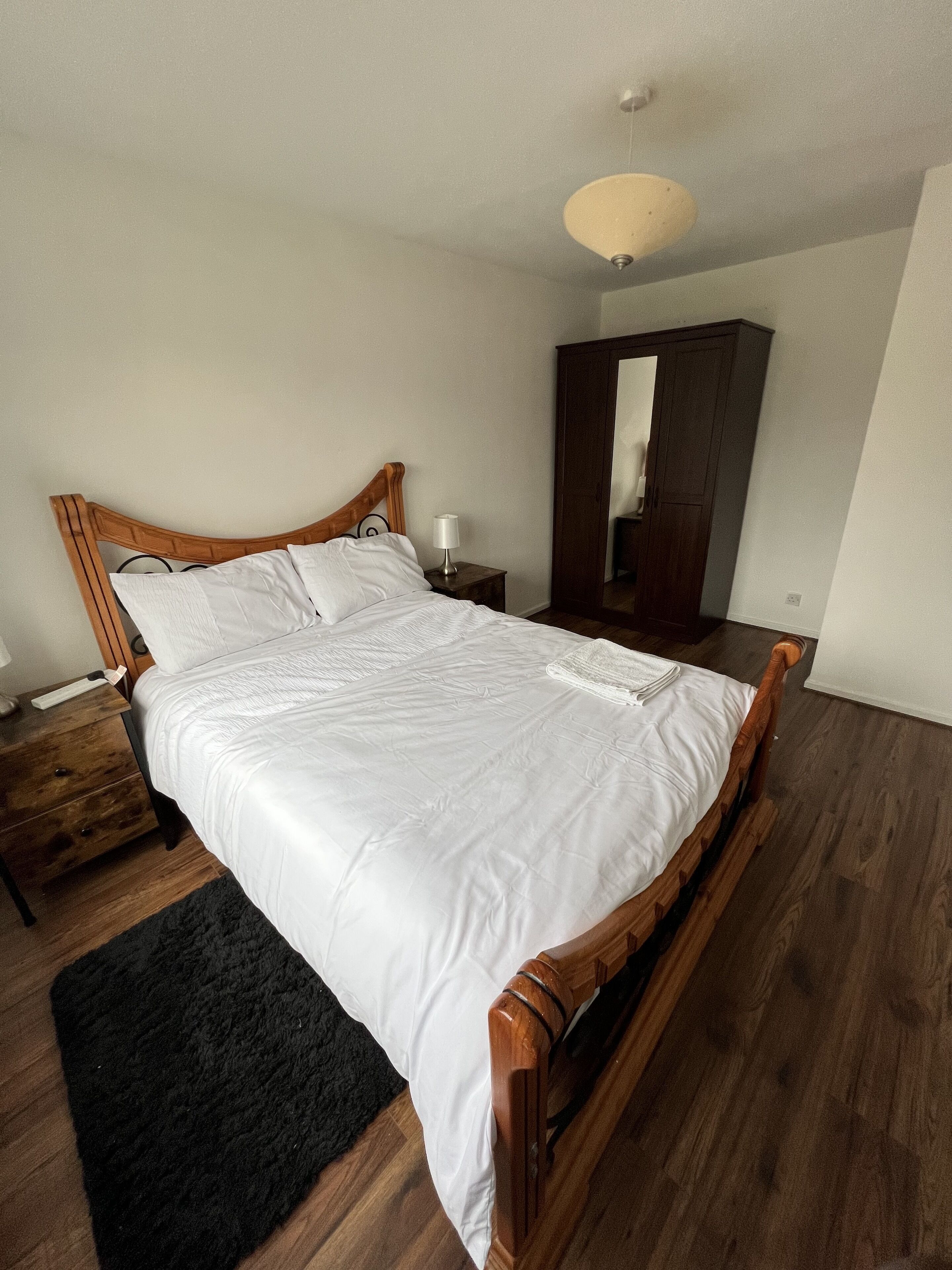 Een net opgemaakt bed met witte lakens, een houten hoofdbord, een nachtkastje met een lamp en een donker houten vloer.