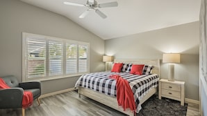 A bedroom with a bed, two bedside tables, a chair, a ceiling fan, and a window with blinds.