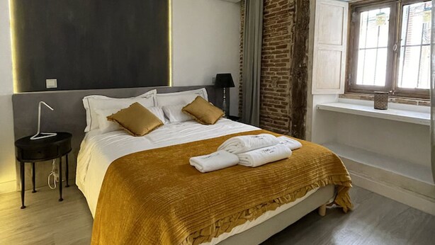 A modern bedroom with a large bed, a bedside table, a window with a view, and a unique ceiling design. - Apartment 2 Rooms MURILLO (Madrid)