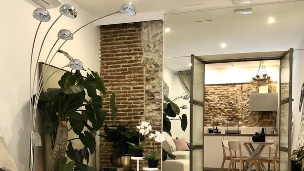 A modern living room with a glass coffee table, white sofa, and large green plants. A kitchen area is visible through an open doorway.