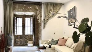 A modern living room with a white sofa, a glass coffee table, and a large plant. There is a rustic stone wall and a large window with curtains.