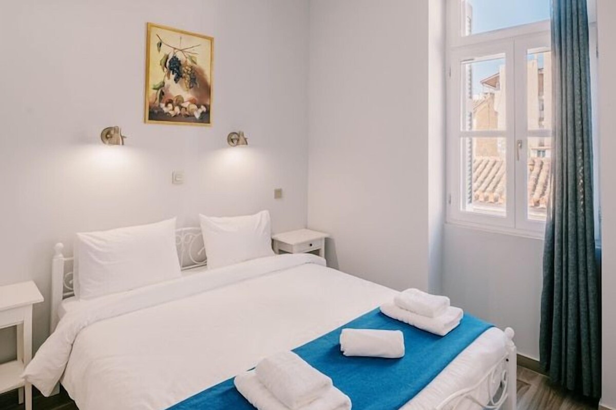 A hotel room with a bed, bedside tables, a chandelier, a window with curtains, and a wall-mounted picture.