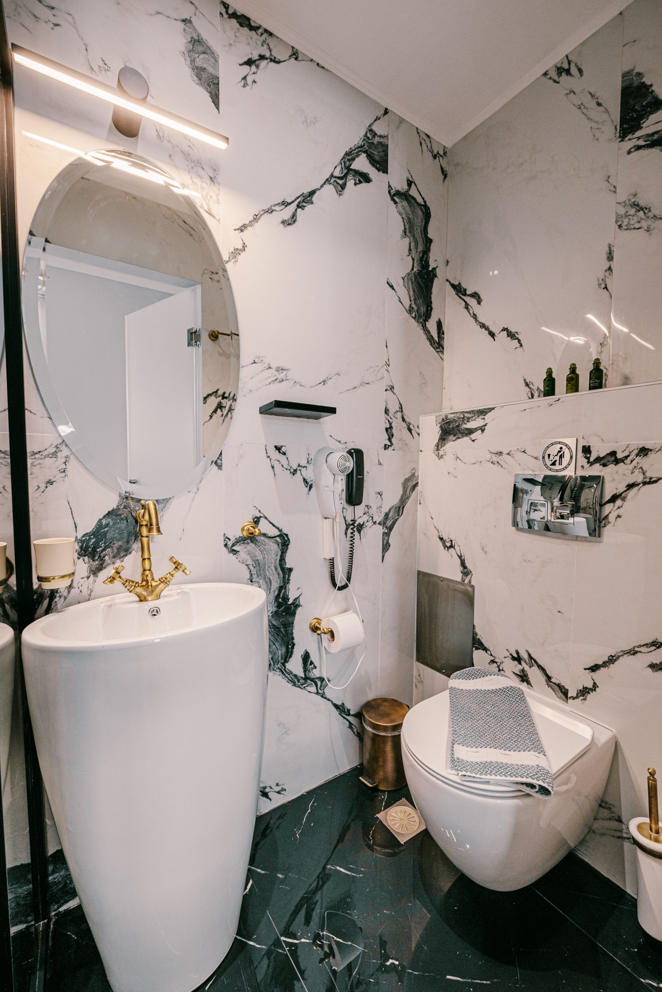A bathroom with a large white sink, a gold faucet, a round mirror, a toilet, and a marble-patterned wall.