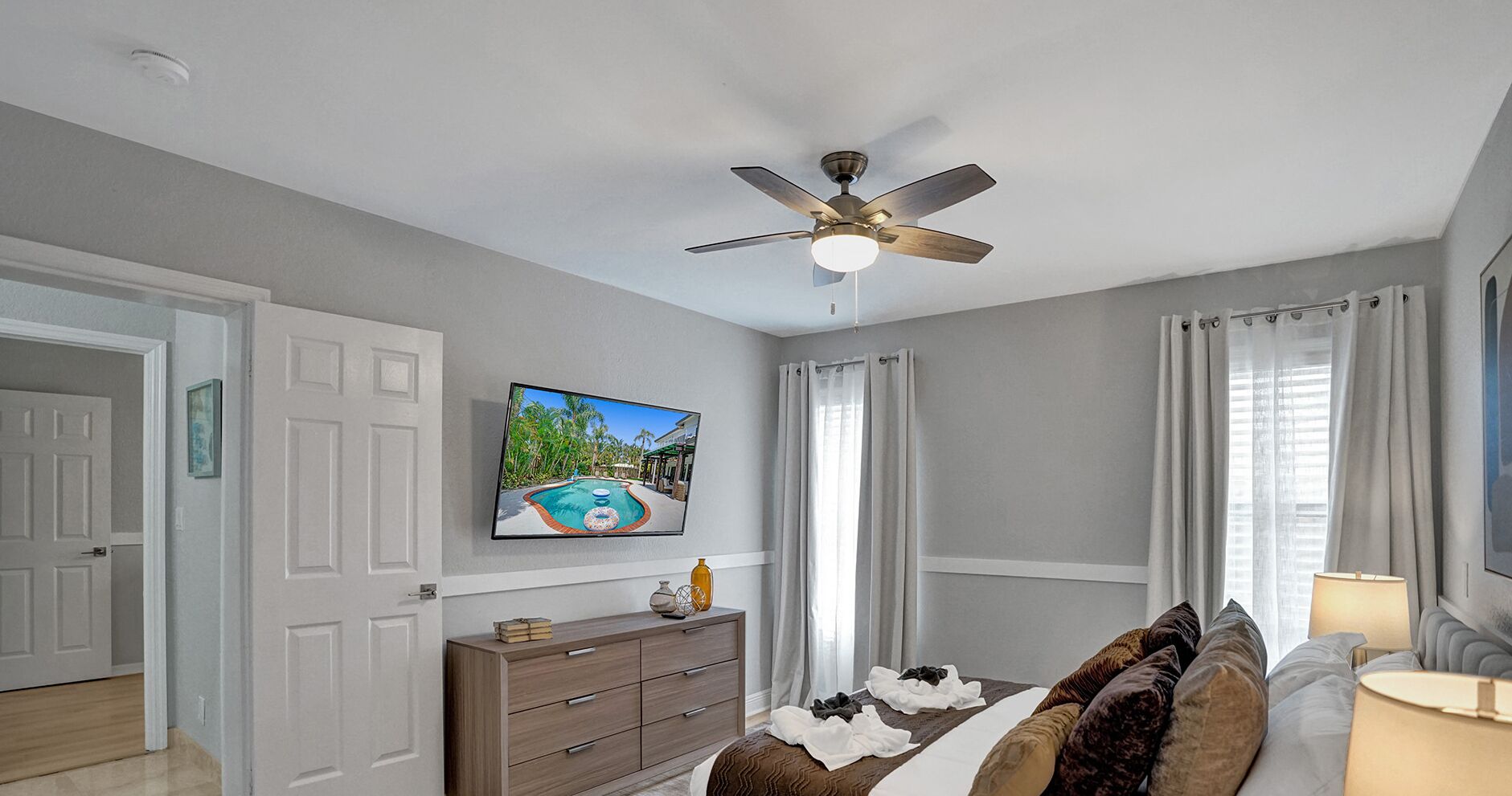 A bedroom with a ceiling fan, a bed with a brown and white patterned bedspread, a dresser, a television on the wall, and a window with curtains.