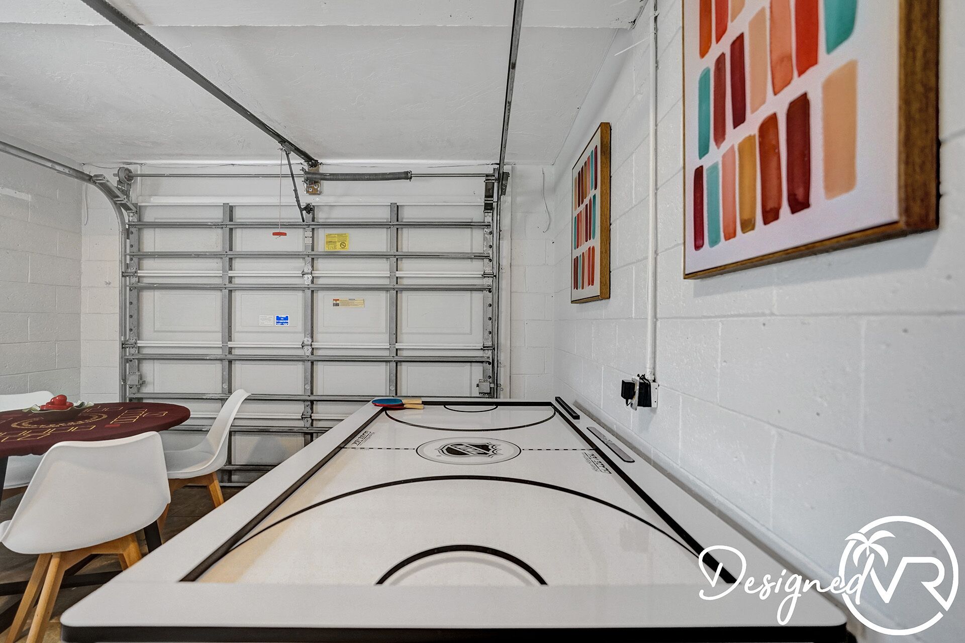 Une pièce avec une table de billard, des chaises, un mur blanc orné d’un tableau coloré et une porte de garage.