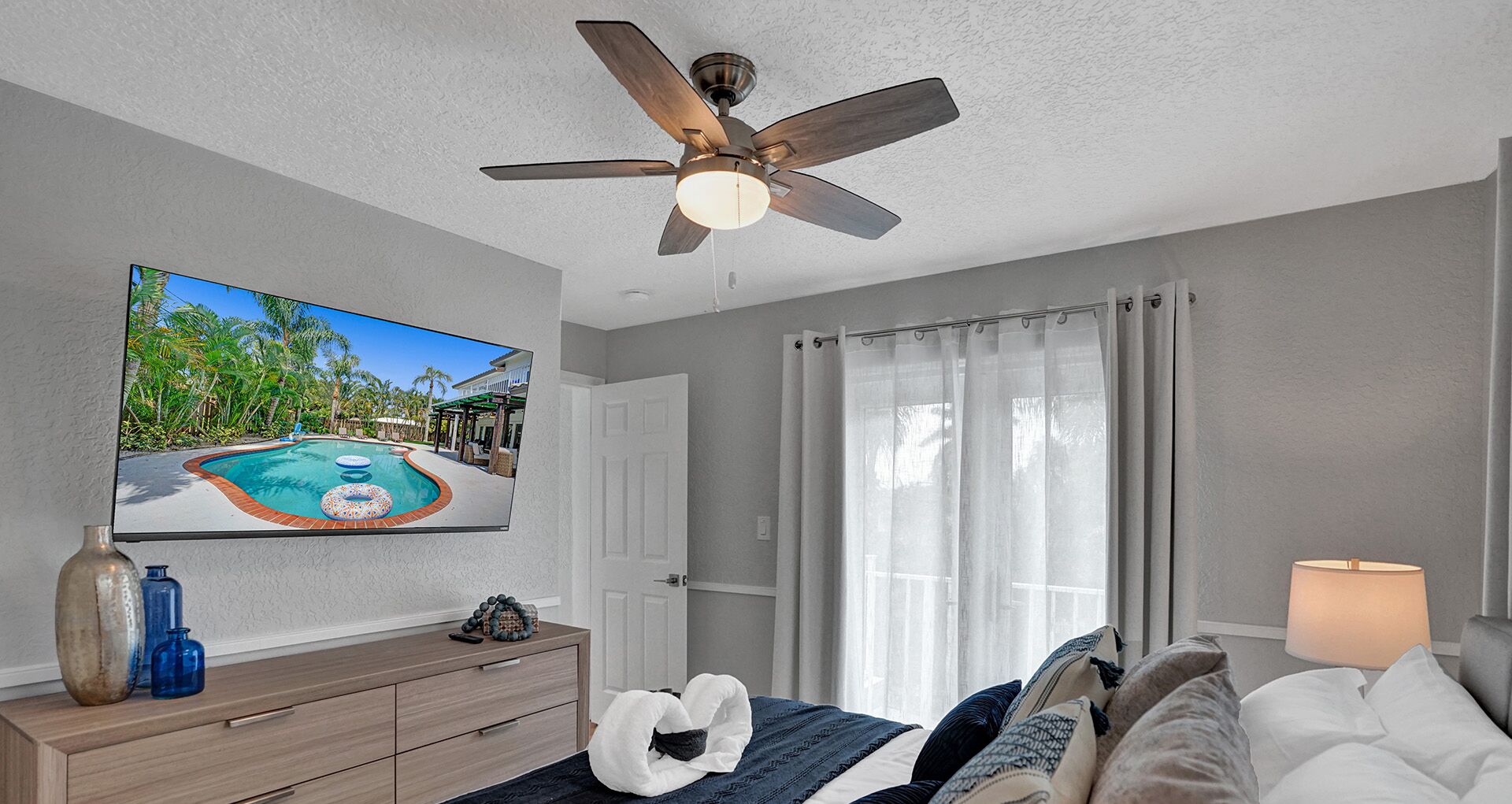 A bedroom with a ceiling fan, a TV displaying a pool scene, a bed with pillows, a bedside table with a vase and remote, and a window with curtains.