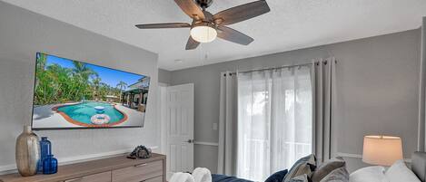 A bedroom with a ceiling fan, a TV displaying a pool scene, a bed with pillows, a bedside table with a vase and remote, and a window with curtains.