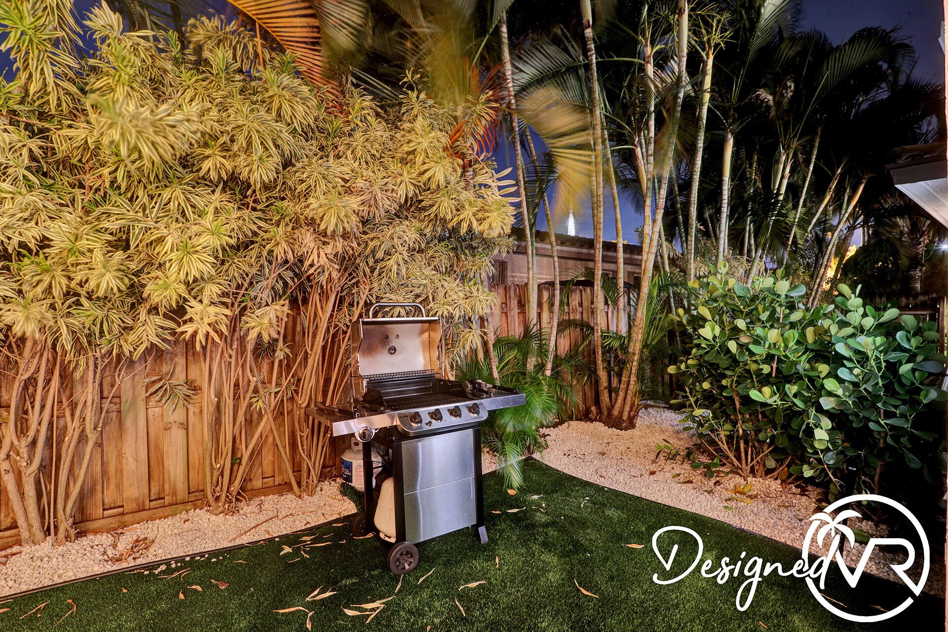 Un jardin avec un barbecue, une clôture en bois et différentes plantes tropicales.
