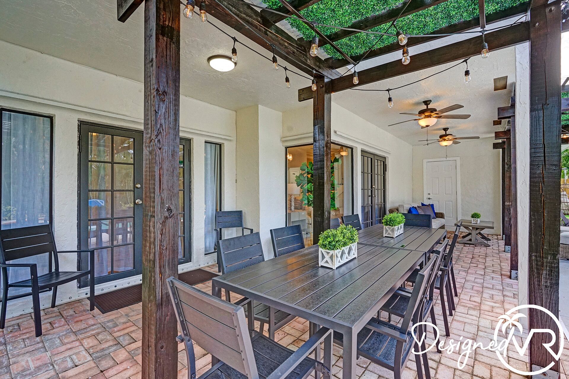 An outdoor patio with a long wooden table and chairs, surrounded by large windows and doors.