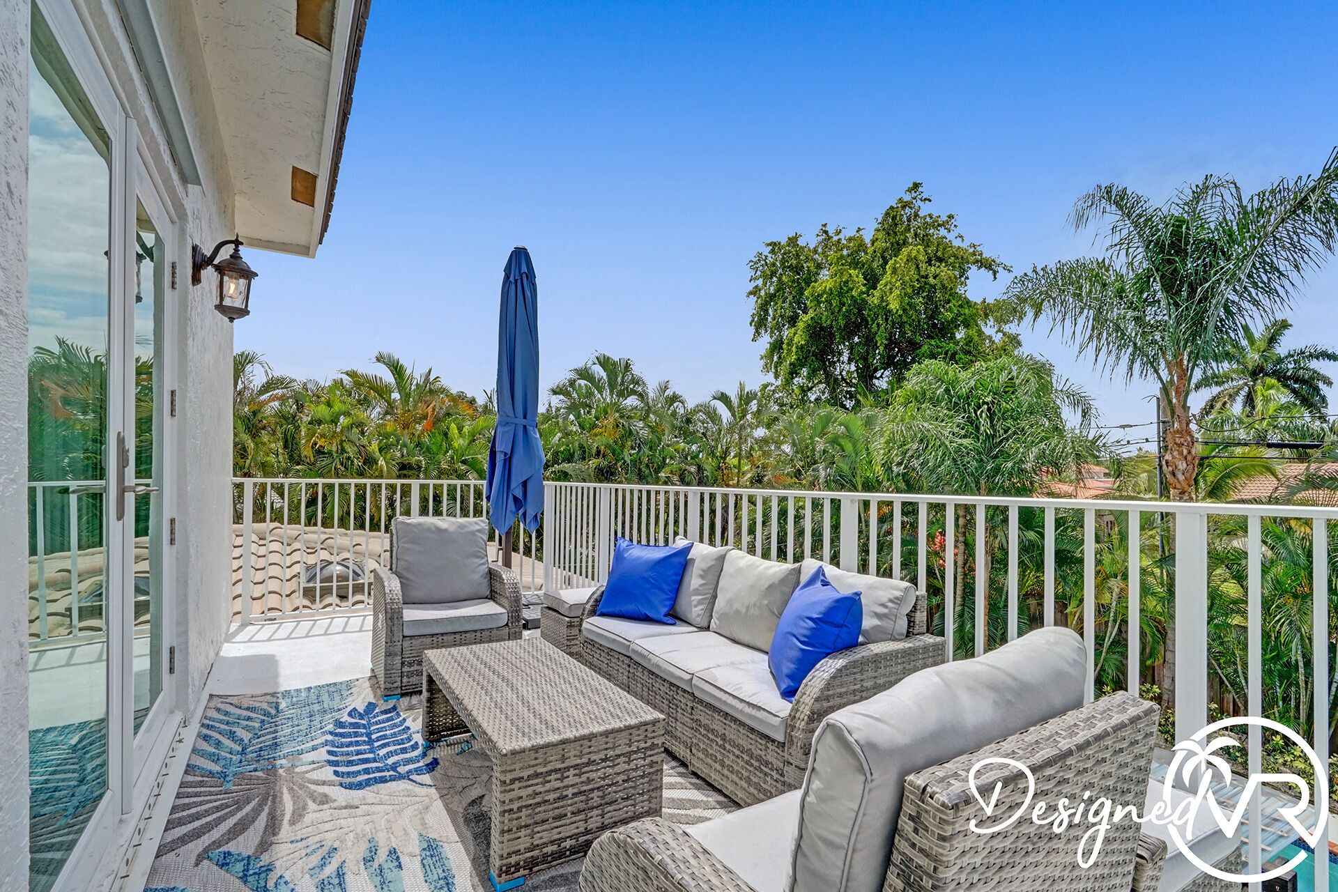 Une terrasse extérieure avec des meubles en osier, un parasol bleu et une vue sur des arbres tropicaux.