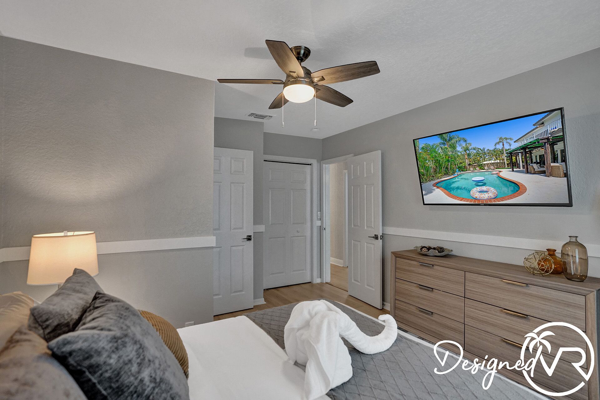 A bedroom with a ceiling fan, a TV displaying a pool scene, a bed with a white comforter, and a wooden dresser.