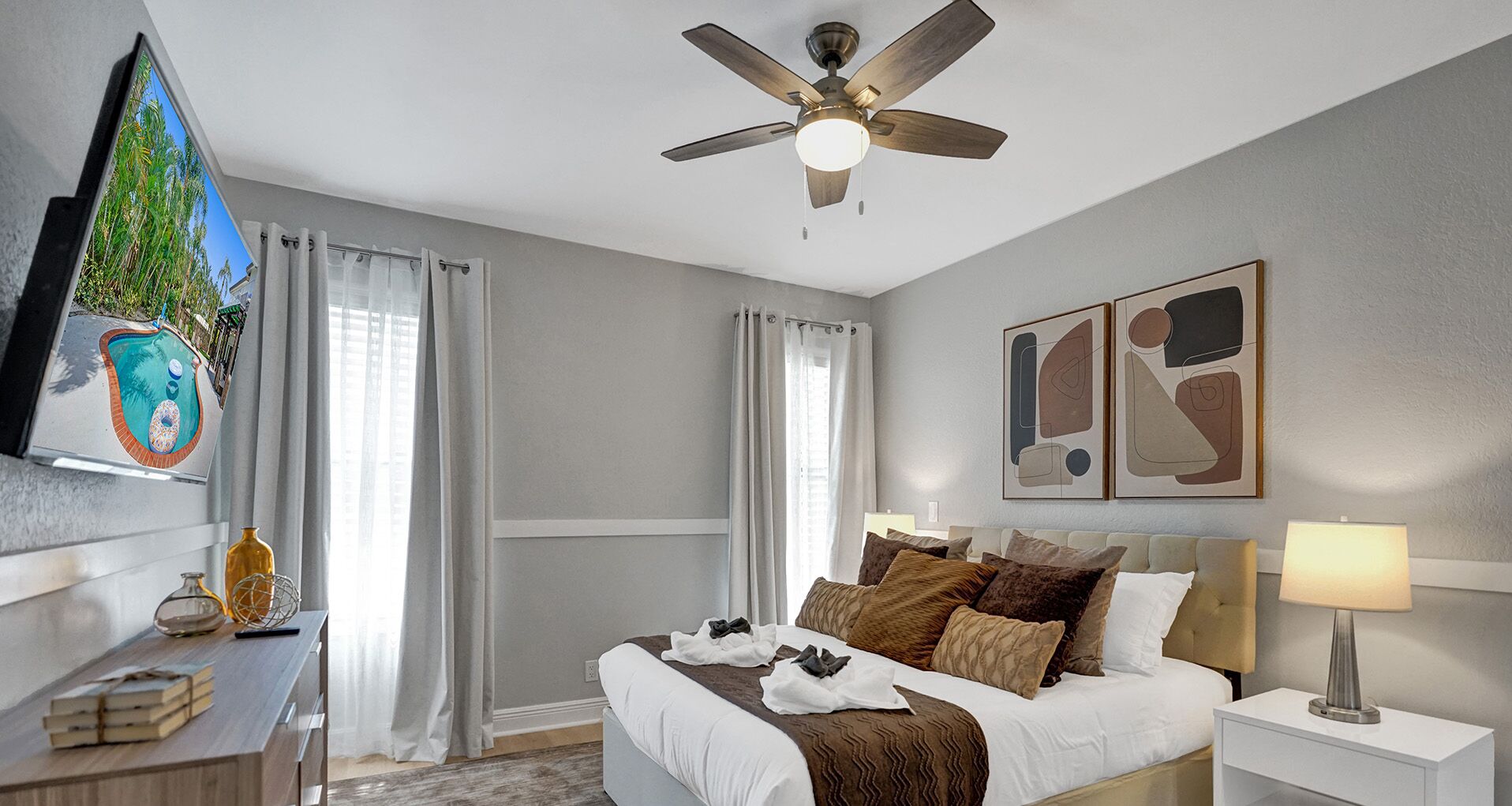 Habitación de hotel con cama, ventilador de techo, mesita de noche con lámpara, un cuadro en la pared y vista a una piscina.