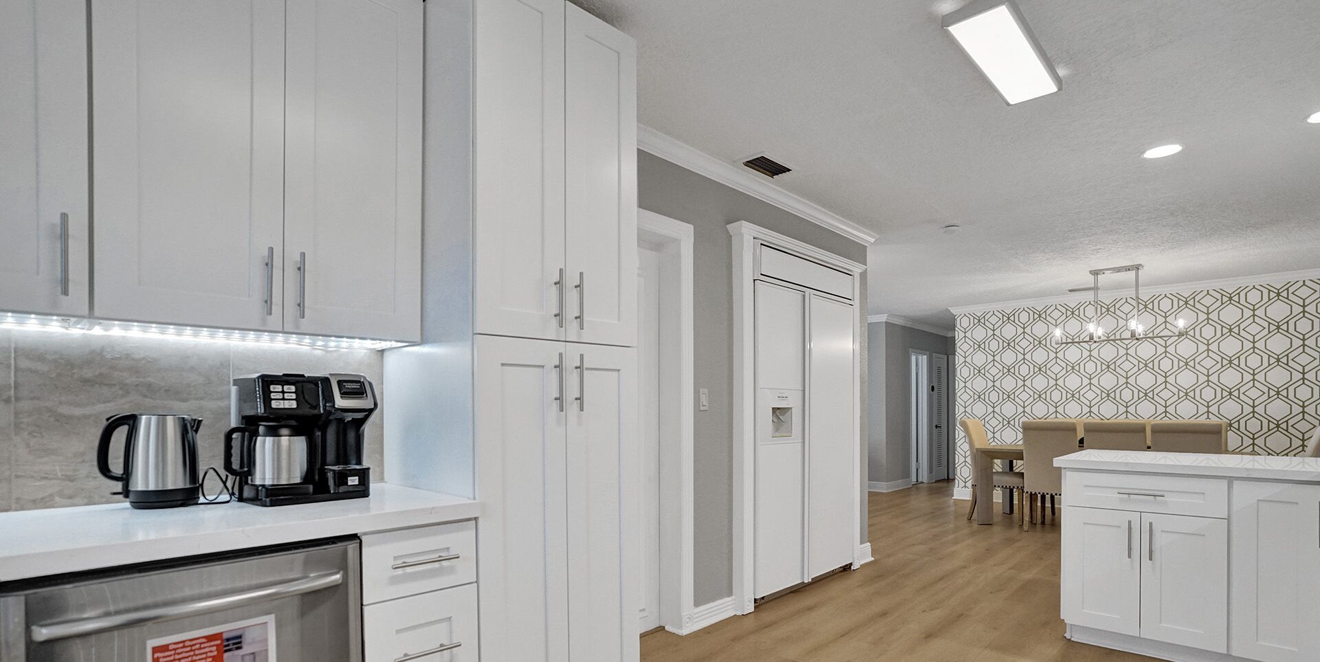 A modern kitchen with white cabinets, a coffee machine, and a dining area with a patterned wallpaper and a wooden table.