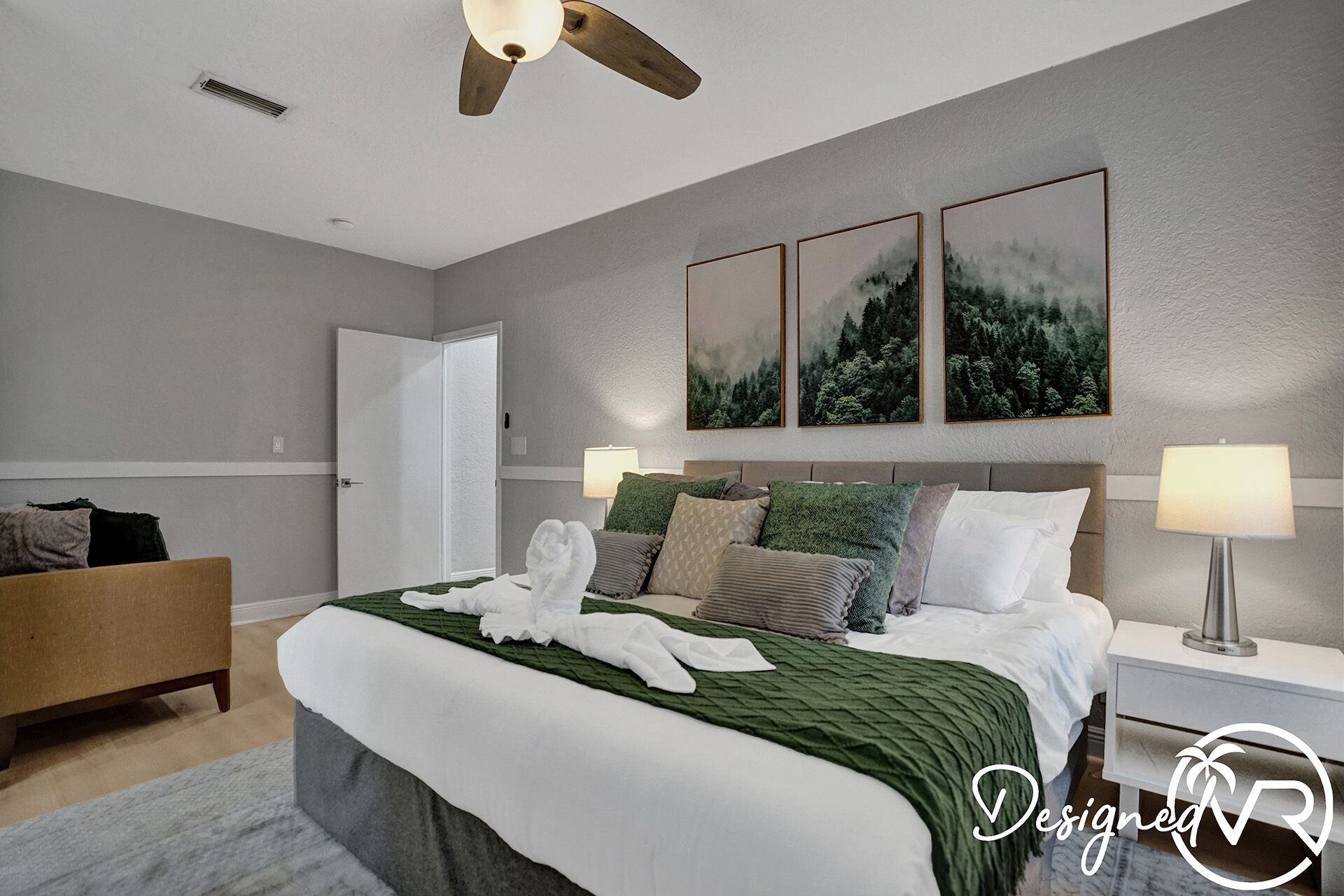 Une chambre à coucher avec un lit, deux lampes de chevet, un ventilateur de plafond, une porte et trois tableaux encadrés accrochés au mur.
