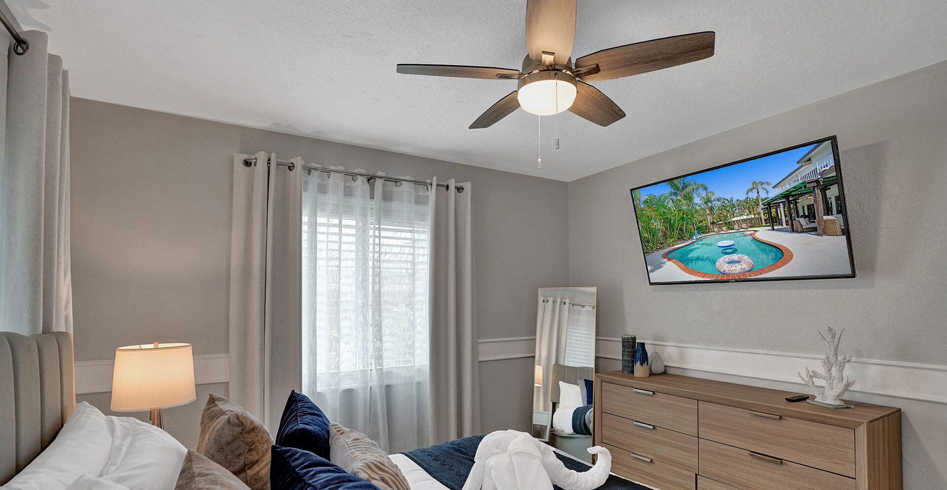 Una habitación de hotel con una cama, un ventilador de techo, un televisor mostrando una piscina, una mesita de noche y un aparador.