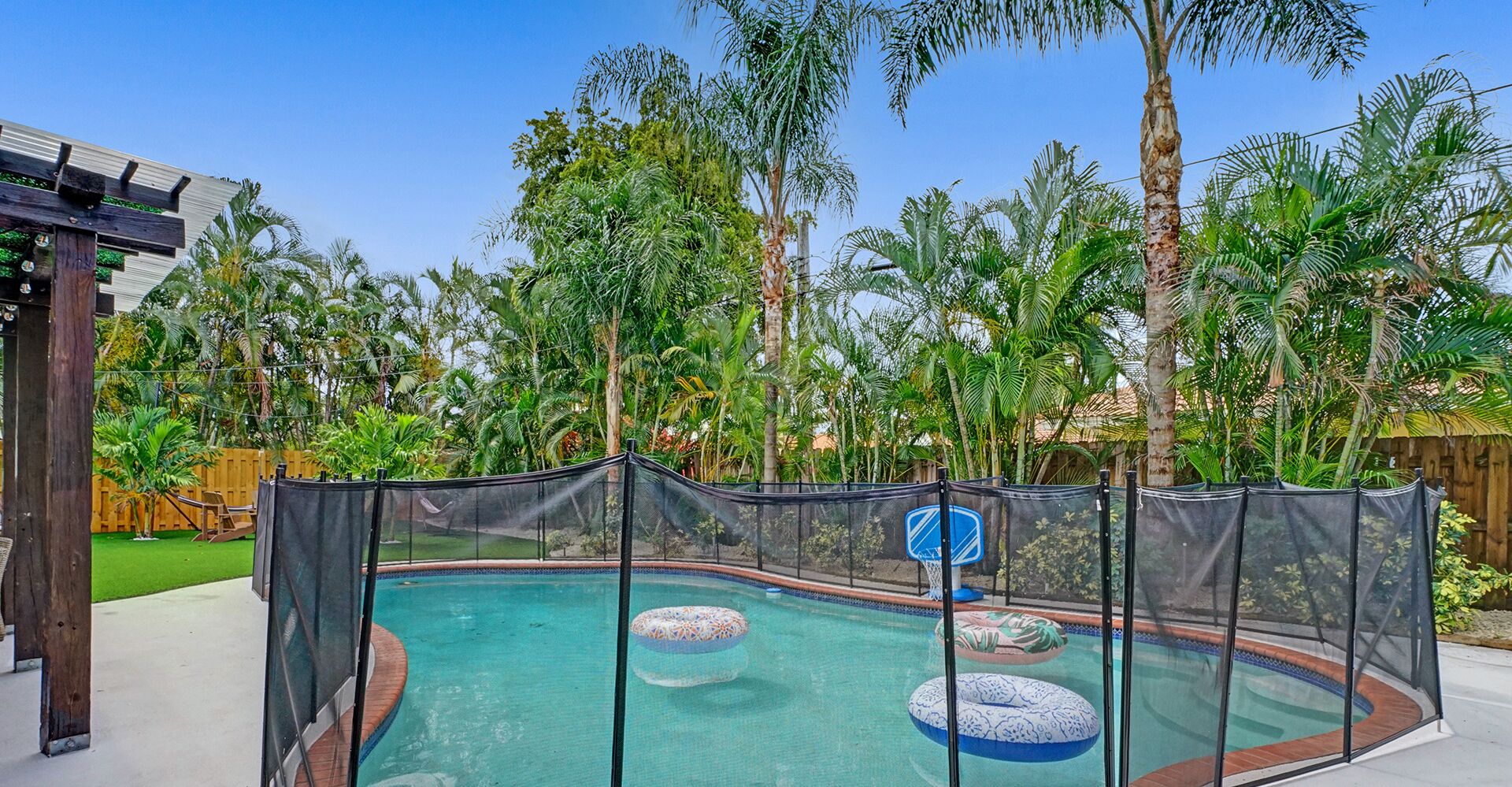 Piscina com cerca de segurança, cercada por palmeiras e um deque de madeira.