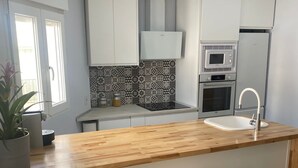 Una cocina moderna con armarios blancos, encimera de madera, microondas integrado, horno y fregadero. Hay dos taburetes de madera y una planta en maceta.