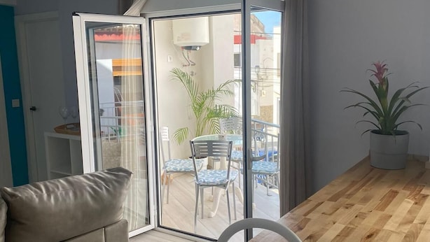 Un salón moderno con suelo de madera, un sofá, un lavamanos y un ventanal corredero que da a un patio exterior con una mesa y sillas.