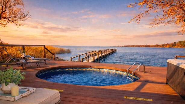 Een houten terras met een bubbelbad, een houten steiger die het water inloopt, en uitzicht op water en lucht.