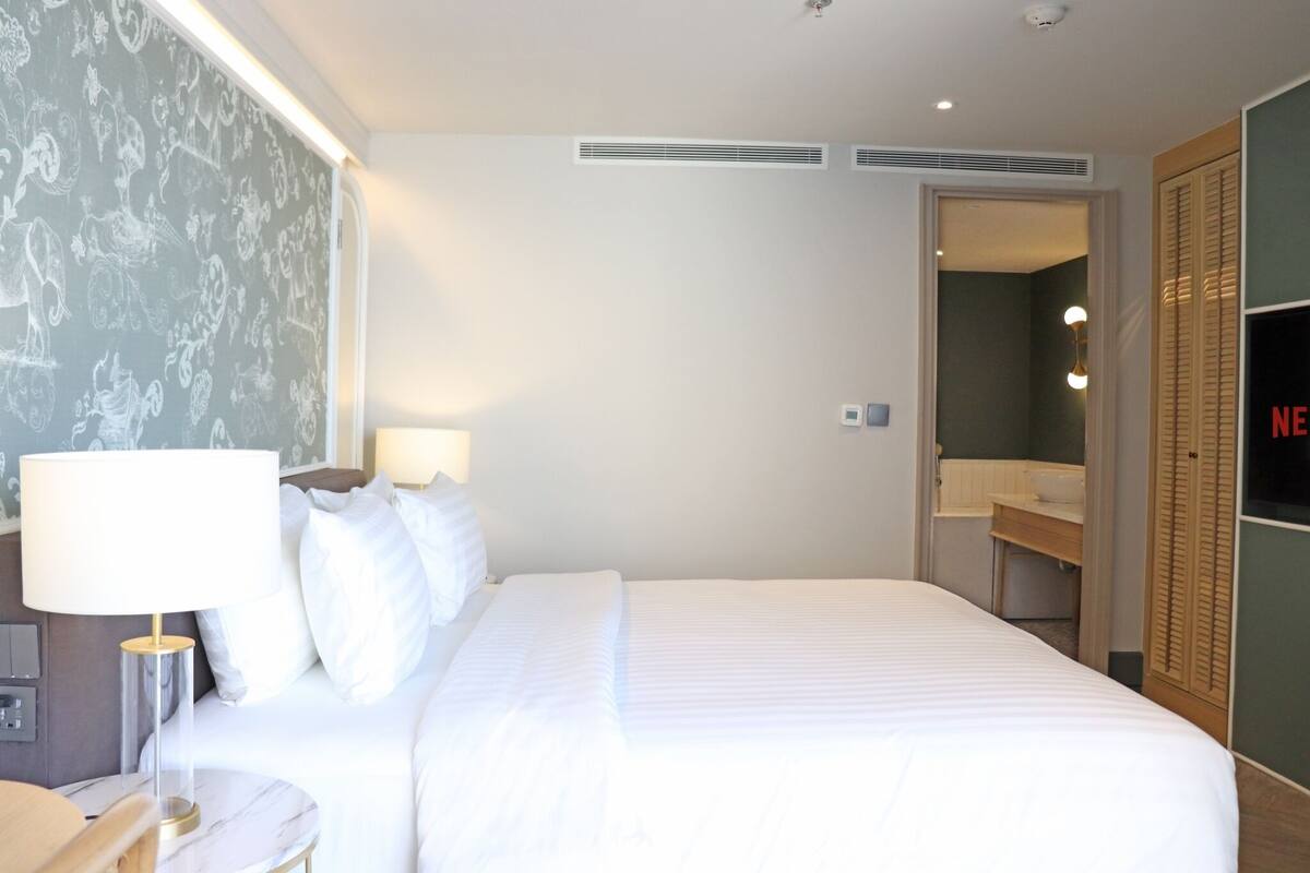 A hotel room with a large bed, a bedside table with a lamp, a wardrobe, and a bathroom visible through an open door.