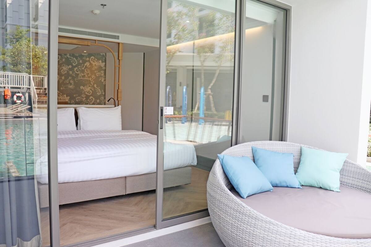 A modern hotel room with a large bed, a sofa with cushions, and a glass wall offering a view of the outside.