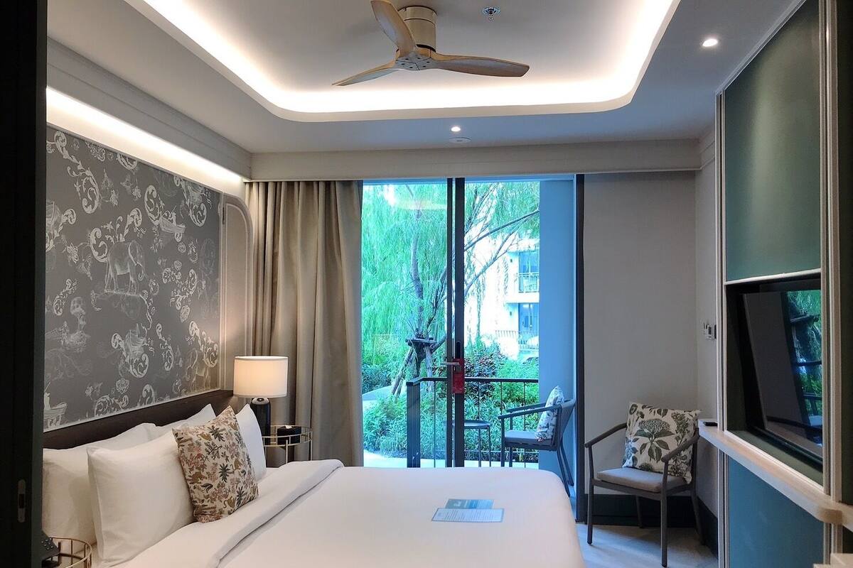 A modern hotel room with a large bed, a desk, a chair, a TV, and a balcony with a view of greenery.