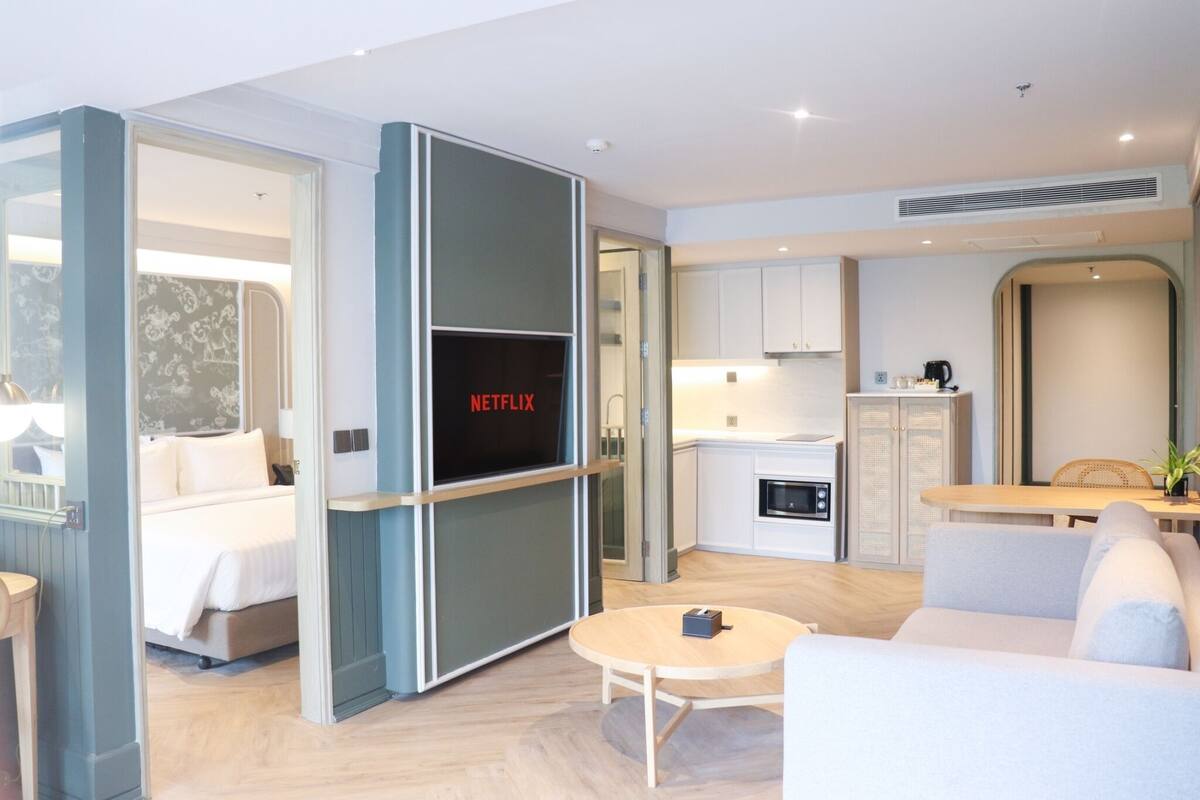 A modern hotel room with a bed, a TV screen displaying 'NETFLIX', a dining area, and a kitchenette.