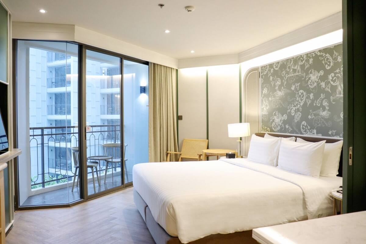 A hotel room with a large bed, a small table, a chair, and a balcony with a railing.