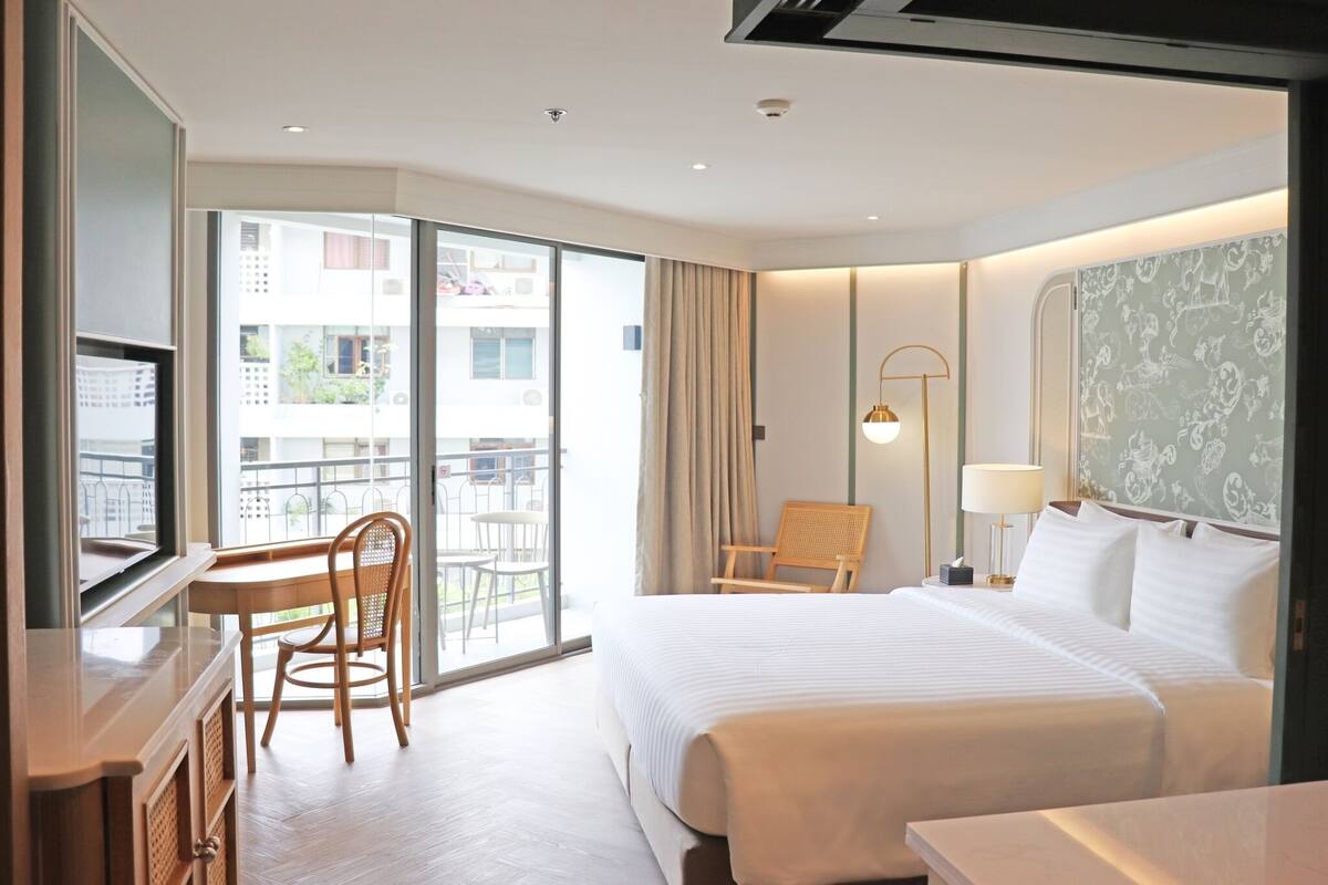 A hotel room with a large bed, a desk with a chair, a balcony with a view of buildings, and a wall with a patterned design.