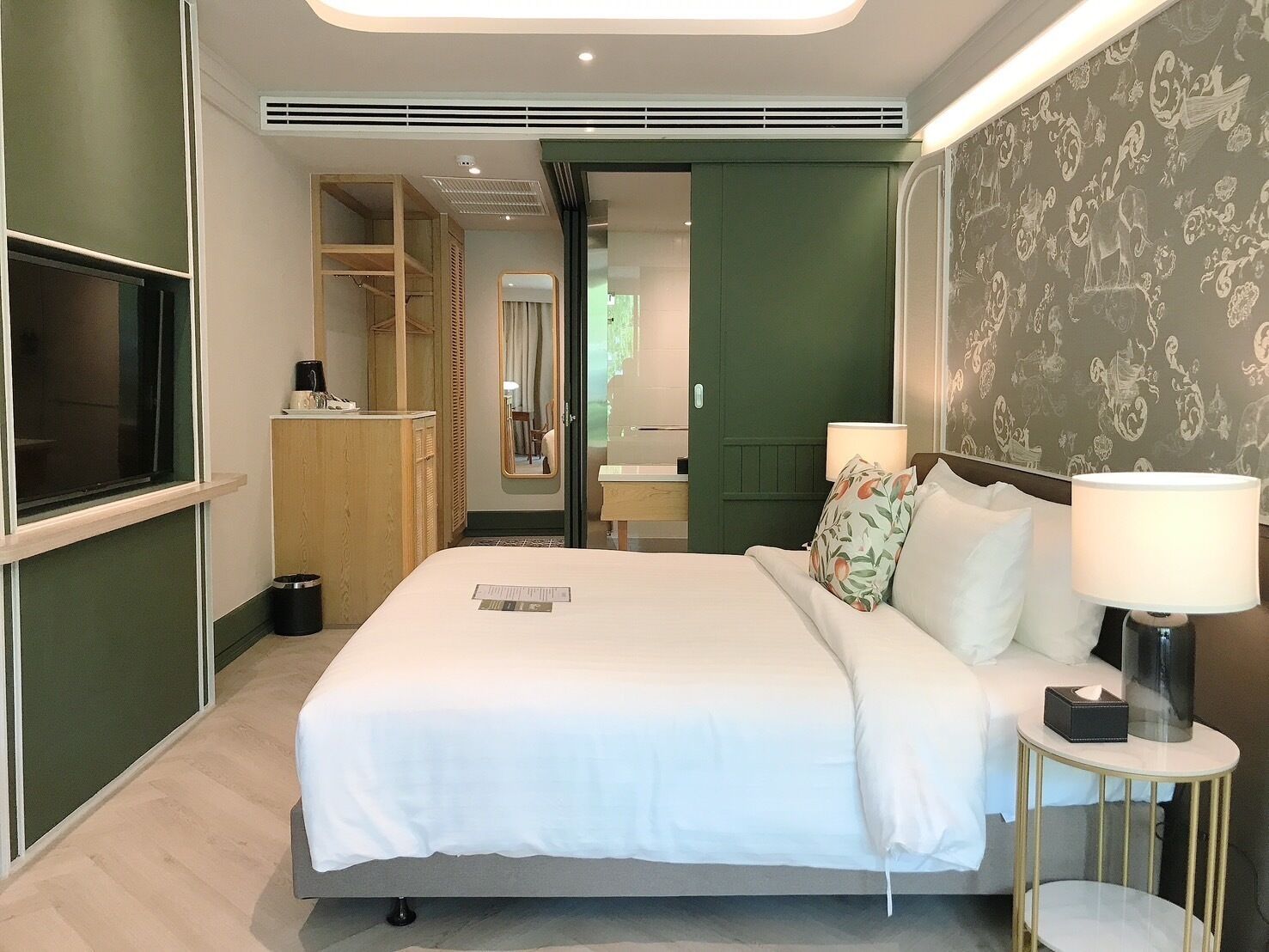 A modern hotel room with a bed, bedside lamps, a TV, a wardrobe, and a bathroom visible through an open door.