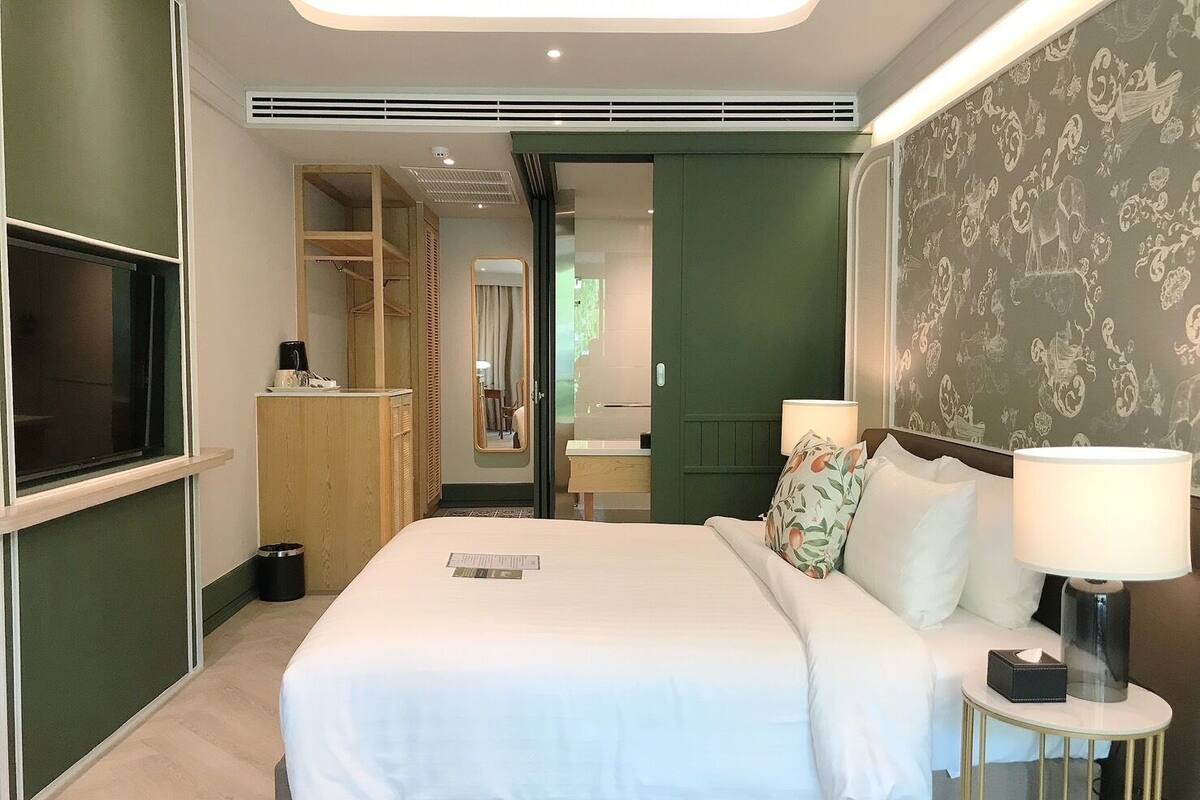 A modern hotel room with a bed, bedside lamps, a TV, a wardrobe, and a bathroom visible through an open door.