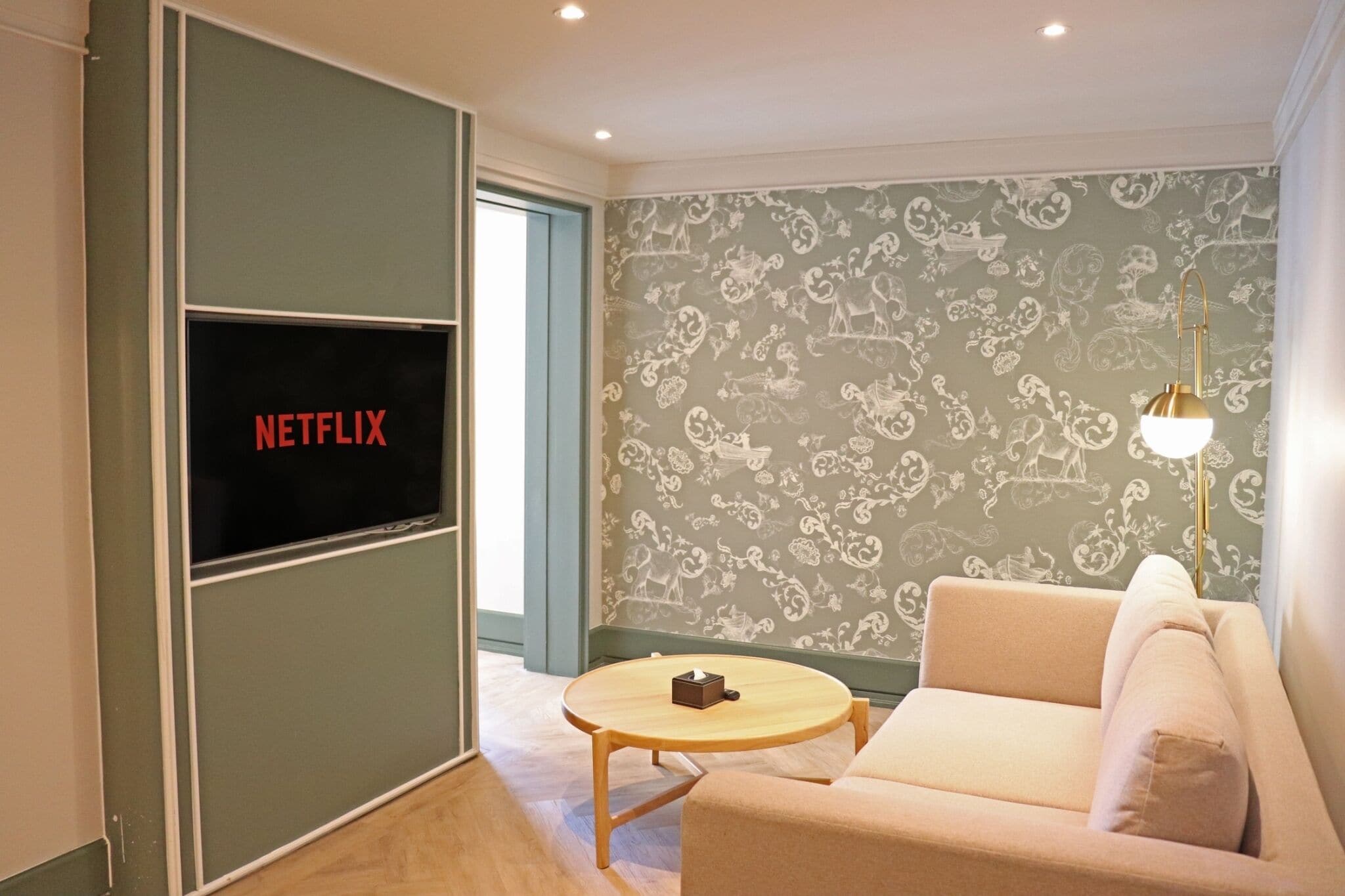 A room with a television displaying 'NETFLIX', a patterned wallpaper, a sofa, a small round table, and a floor lamp.